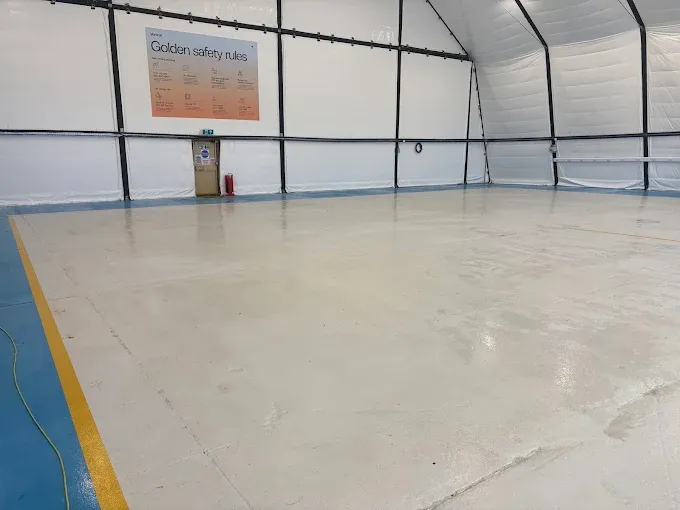 Empty indoor training hall with a white glossy floor, blue trim along the edges, and a 'Golden safety rules' poster on the far wall beside a trash bin.