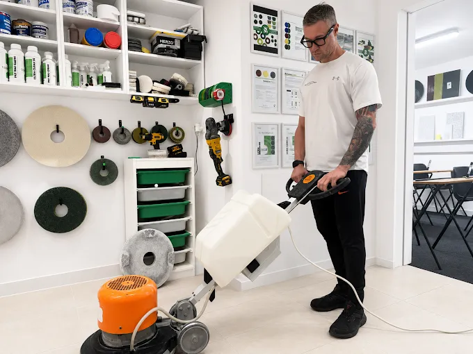 Man in a white t-shirt using a floor polishing/sanding machine in a workshop with shelves of discs and tools on the wall behind him.