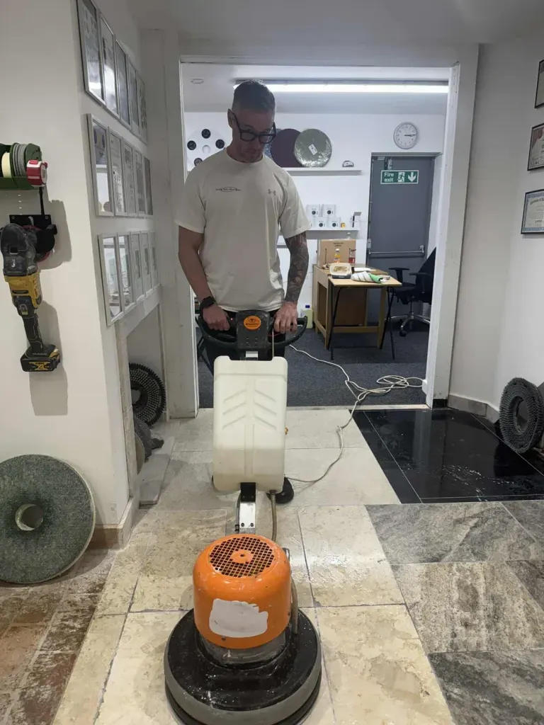 Man operating a floor buffing machine on a travertine floor in a workshop, with polishing pads and discs on the wall.