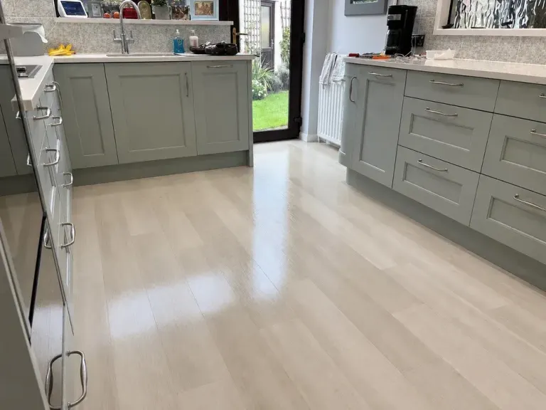 Glossy light wood-effect vinyl plank flooring in a kitchen with sage green cabinetry and garden doors.