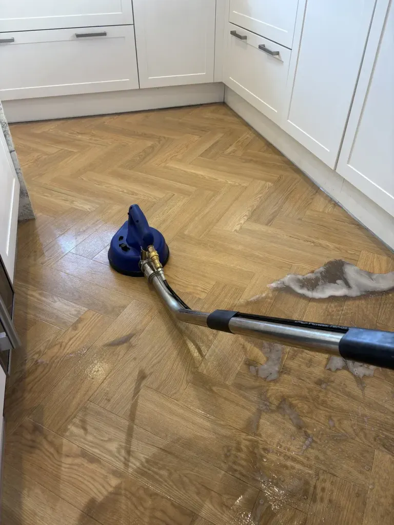 Blue extraction tool head cleaning grime from a herringbone wood-effect kitchen floor, hose trailing across the boards.