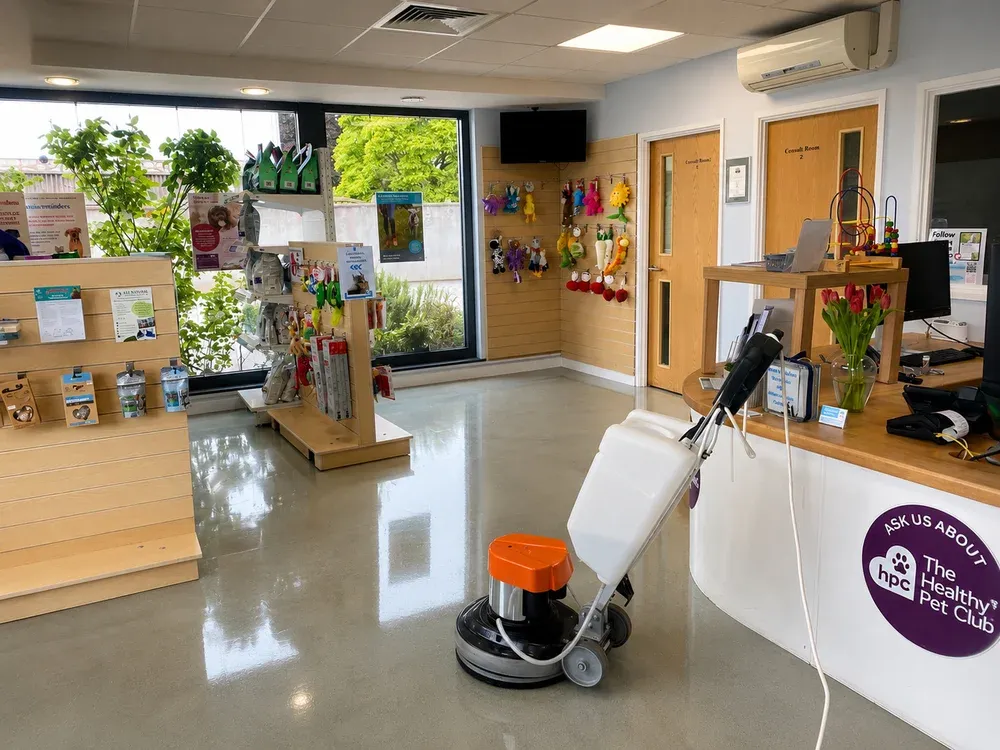 Orange floor buffing machine polishing a veterinary practice reception floor, with pet toys and shelving nearby.