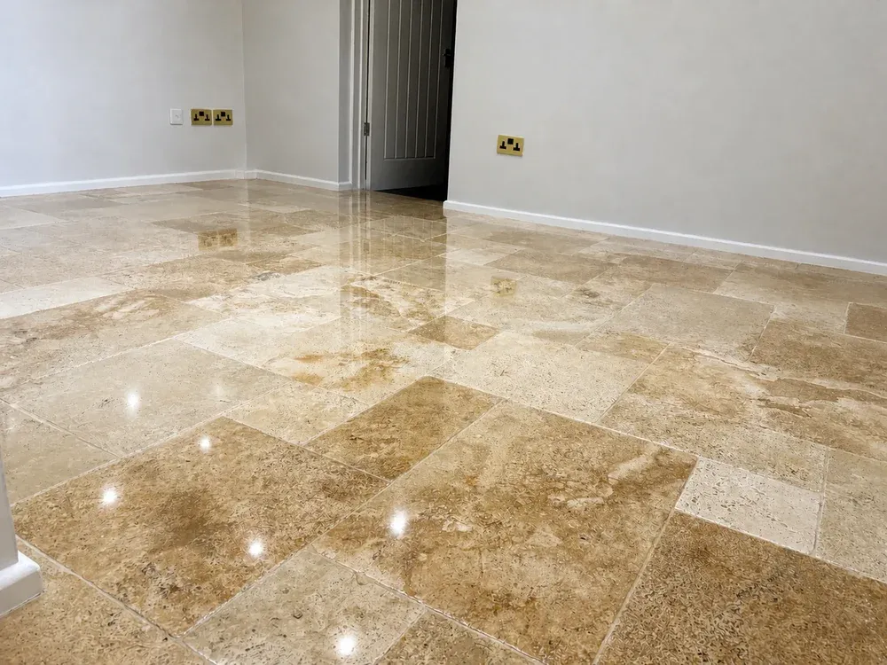 Glossy polished travertine floor in an empty room, reflecting the doorway and brass sockets on the wall.