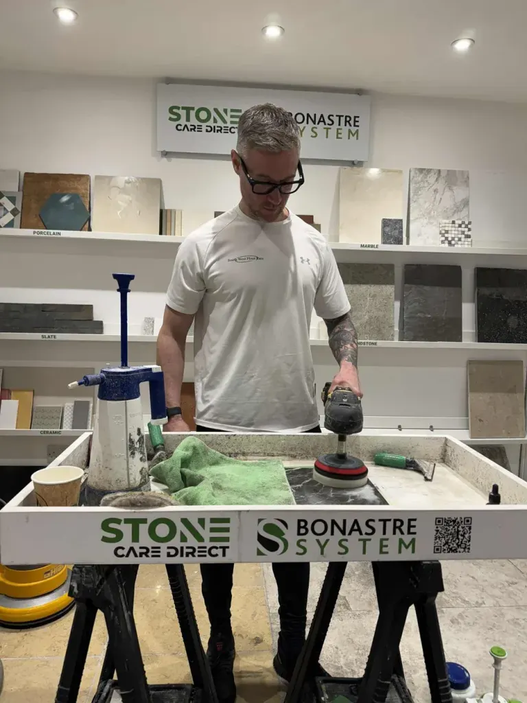 Man polishing a small stone sample slab at a training bench, surrounded by marble, slate and ceramic sample tiles.