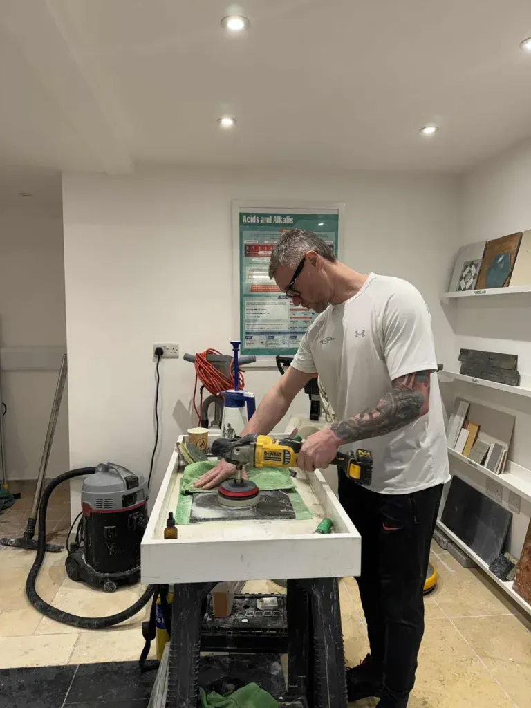 Technician using a handheld polisher on a stone sample at a workbench, surrounded by tile samples in the workshop.